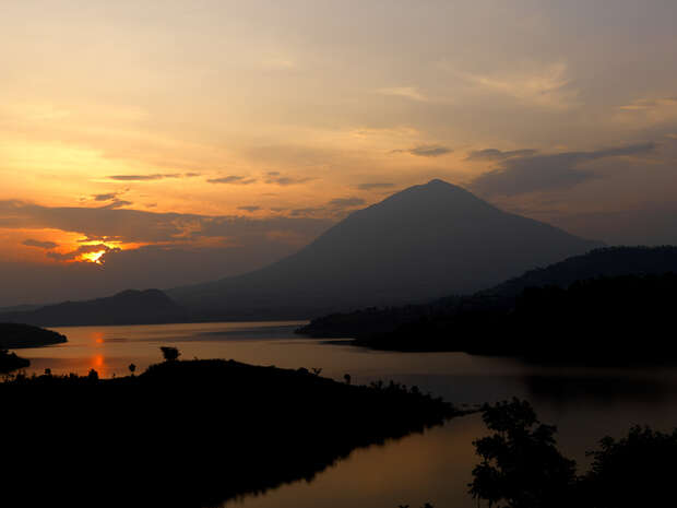 A mountain stands silhouetted against an orange and yellow sunset sky, with calm water reflecting the warm colors, surrounded by dark tree-covered hills.