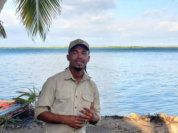 A person stands near a calm, palm-lined beach with blue water and partly cloudy sky. They wear a beige shirt and cap, gesturing with one hand while looking towards the camera.