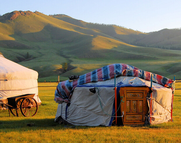 A traditional yurt stands on a grassy plain, illuminated by sunset, with verdant hills in the background. Adjacent is a cart with a canvas cover, enhancing the rustic scene.