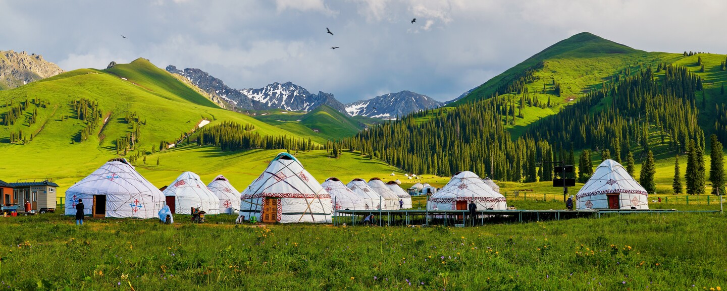 Yurts sit on a grassy plain, framed by green, pine-covered hills and snowy mountains under a cloudy sky, with a few people nearby and birds flying overhead.