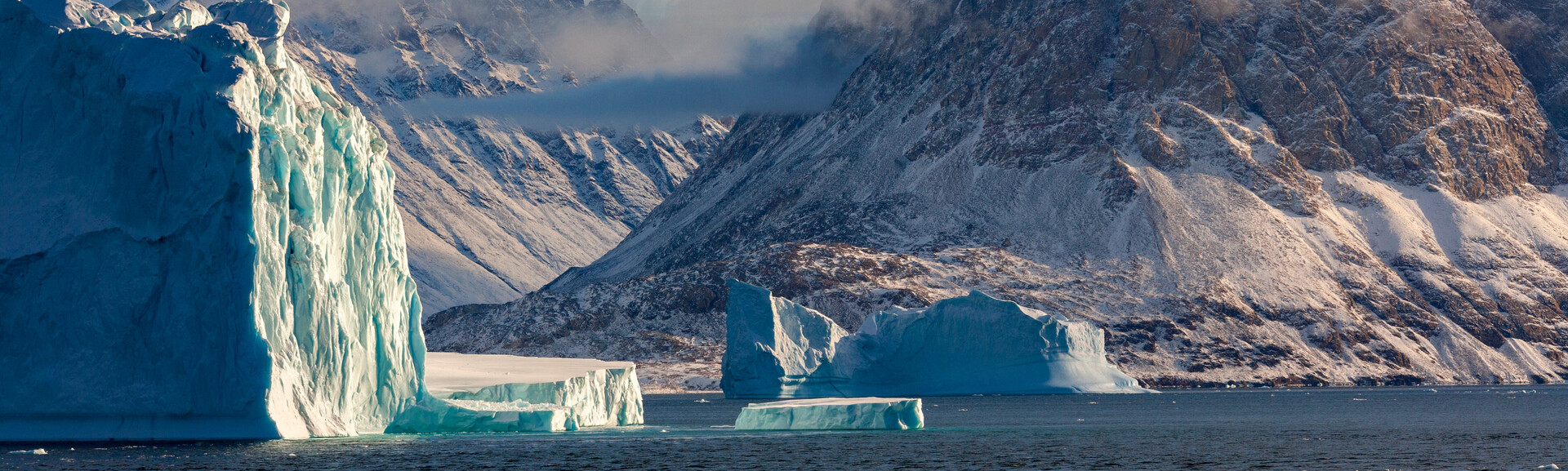 Icebergs float in calm water near snowy, rugged cliffs. The scene is bathed in soft light, with clouds partially covering the mountains in the background, creating a serene Arctic landscape.