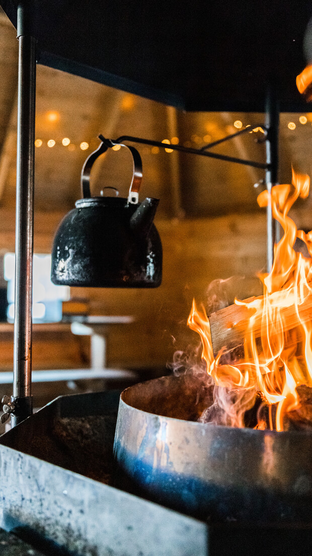 A black kettle hangs over bright flames on a metal stand, inside a dimly-lit wooden room decorated with string lights.