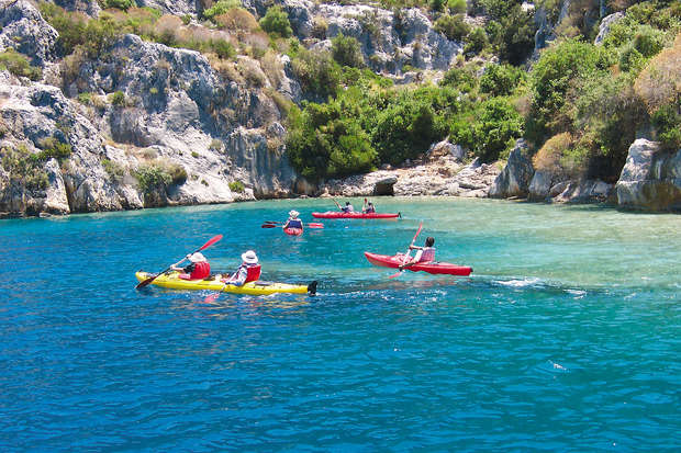 Kayaks glide across vibrant blue water, navigated by people wearing hats and paddling. Rocky cliffs and lush greenery form a scenic backdrop under a clear sky.