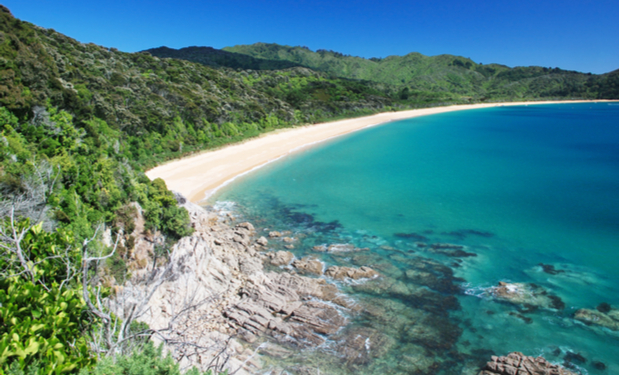 Turquoise ocean waters gently lap a sandy crescent beach, bordered by rocky outcrops and lush green forested hills under a clear blue sky.