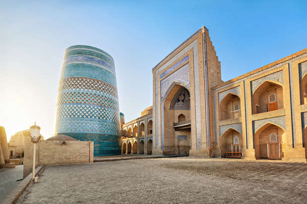 A tall, cylindrical, blue-tiled tower stands beside a large, ornately decorated building with archways; both set in a paved courtyard under a clear blue sky.