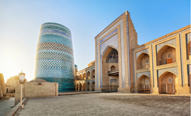 A large, ornate minaret with blue and turquoise tiles stands beside a historic brick building featuring arched doorways and intricate patterns, set under a clear blue sky in a sunlit courtyard.