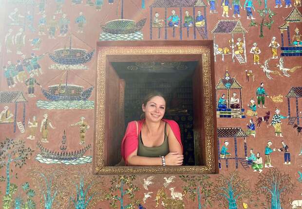 A person leans through an ornately framed window, smiling. The background features intricate, colorful wall art depicting boats, people, and animals in a vibrant, detailed scene.