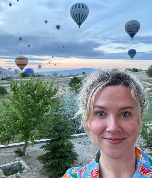 A person smiling with multiple hot air balloons floating in the sky at sunrise, set against a landscape of trees and open fields.