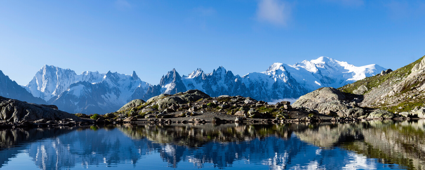 Snow-capped mountains reflect in a clear, tranquil lake, framed by rocky terrain under a bright blue sky.
