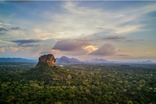 A large rock formation rises amidst a vast, dense forest. The sky above is filled with soft clouds and hues of sunset, with distant mountains on the horizon.