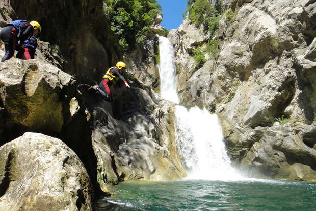 A person wearing a helmet and wetsuit jumps from a rocky ledge into a turquoise pool below a tall, cascading waterfall surrounded by steep, green-covered cliffs.