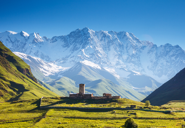 A stone tower stands on a sunlit grassy plain, framed by a backdrop of dramatic snow-covered mountains under a clear blue sky.