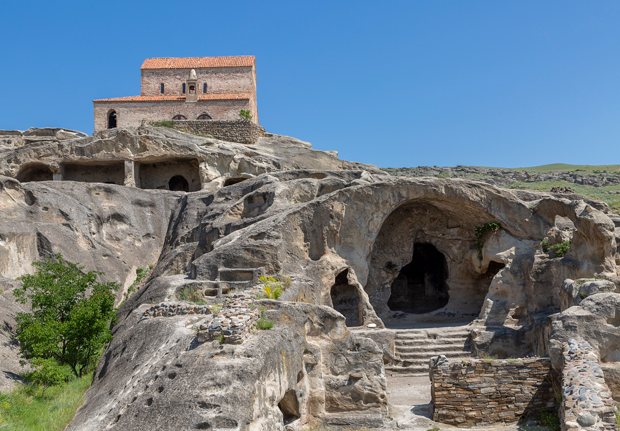 A historic stone building with a red-tiled roof sits atop rocky cliffs featuring cave-like openings, stairways, and scattered vegetation under a clear blue sky.