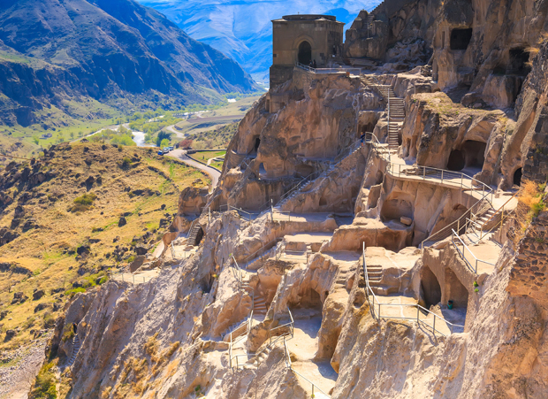 Rock-cut caves form a complex of stairways and passageways. Structures are embedded in a steep mountainside, overlooking a valley with winding roads and distant mountains under a clear blue sky.