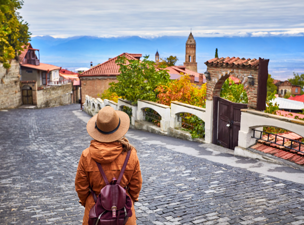A person in a hat and orange coat stands on a cobblestone street, viewing a village with red rooftops and church tower amid autumn trees, under a cloudy sky.
