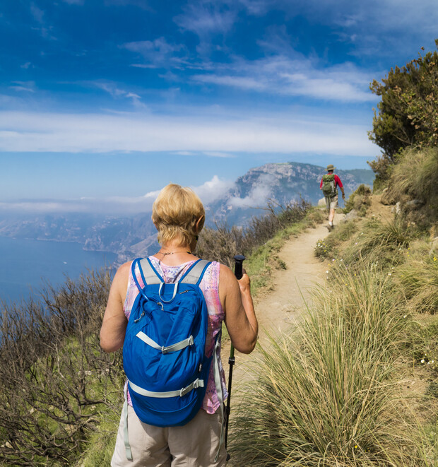 A person with a backpack walks on a narrow mountain trail, using trekking poles. Another hiker is ahead, surrounded by grass and shrubs, with a scenic mountain view in the distance.