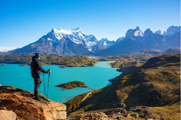 A person stands on a rocky ledge holding trekking poles, overlooking a turquoise lake with snow-capped mountains in the distance under a clear blue sky.