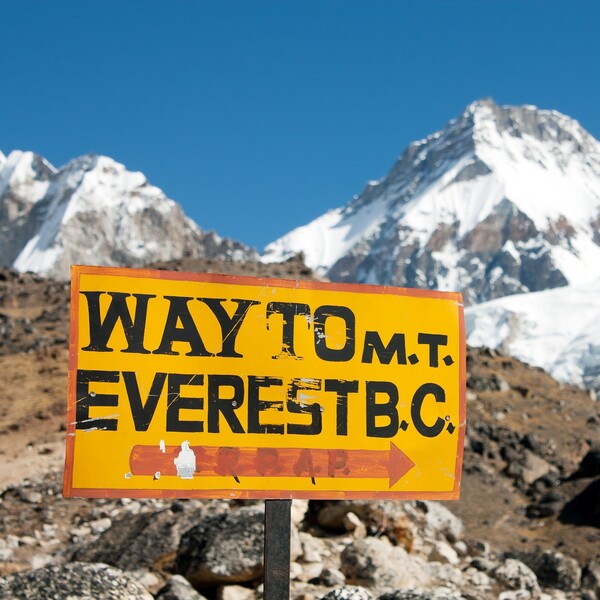 Sign pointing towards Mount Everest Base Camp stands with snow-covered mountains in the background. Text reads: "WAY TO M.T. EVEREST B.C." with an arrow pointing right.