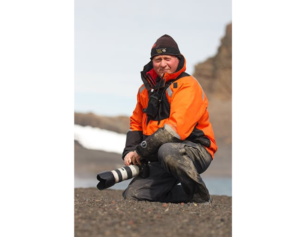 A person in an orange jacket kneels on rocky terrain, holding a camera with a telephoto lens, while a blurred, icy landscape and water provide the background.