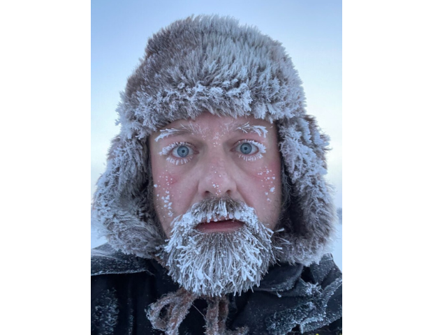 A person with frost-covered eyebrows and beard stands outside, wearing a thick fur hat and winter clothing, surrounded by a cold, icy environment.