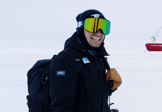 A person wearing dark winter gear and reflective goggles smiles while holding a glove on an icy landscape, with a red ship and crane visible in the distance.