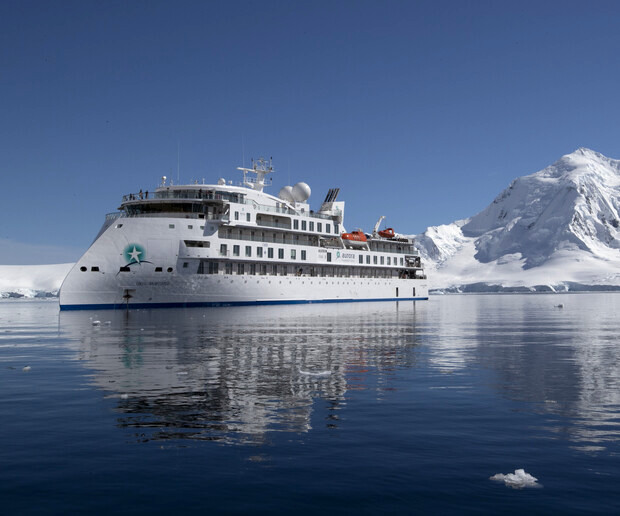A white cruise ship with deck equipment and lifeboats sails calmly on reflective icy waters, surrounded by snow-capped mountains under a clear blue sky.