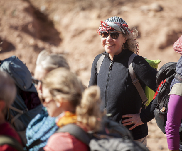 A person wearing sunglasses and a checkered headscarf smiles with a backpack on, surrounded by others in hiking gear, in a sunny, rocky outdoor setting.