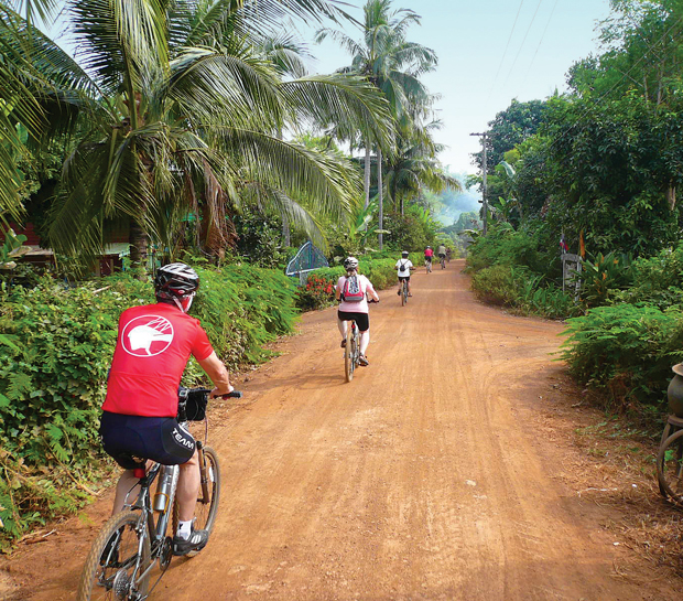 Cyclists ride along a dirt path surrounded by lush greenery and palm trees, with one cyclist wearing a red jersey in the foreground. The road is flanked by dense vegetation.