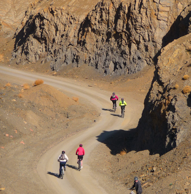 Cyclists ride along a winding dirt path surrounded by steep, rugged cliffs. The rocky landscape is barren, with sparse vegetation, creating a challenging outdoor environment.