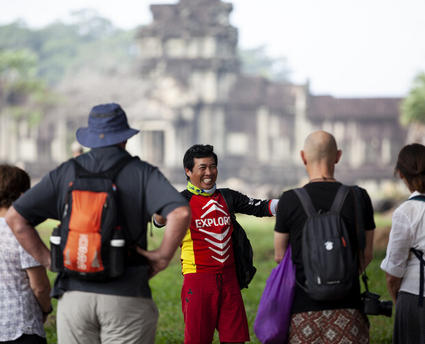 A person in a red "EXPLORE" outfit enthusiastically gestures while speaking to a group of tourists. The background features an ancient stone temple and lush greenery.