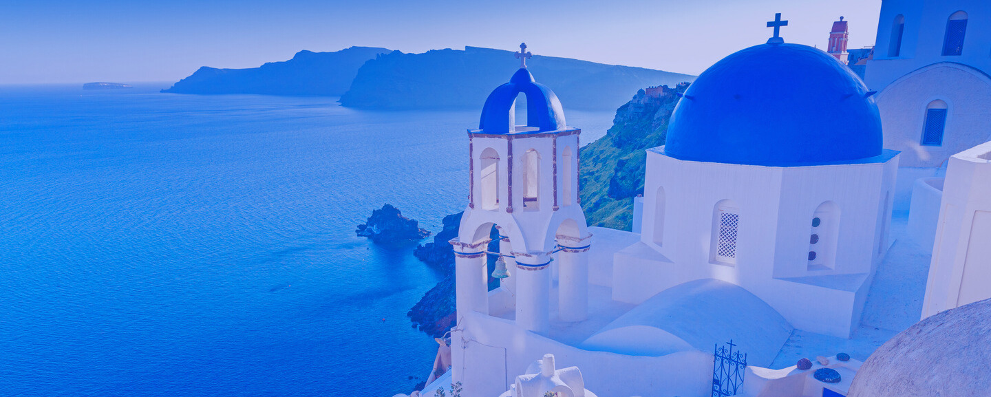 White buildings with blue domes stand on a clifftop overlooking a vast blue sea, with distant islands and a mountainous backdrop under a clear sky.