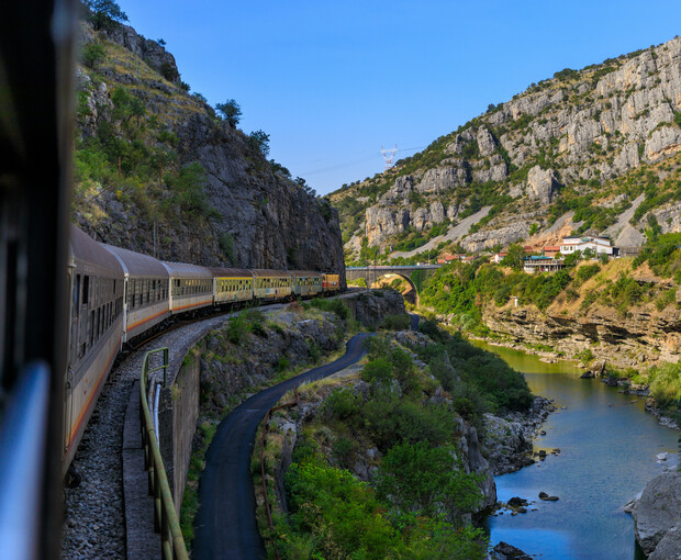 A train curves along a track through a picturesque canyon, bordered by rocky cliffs and a small, flowing river under a bright blue sky.