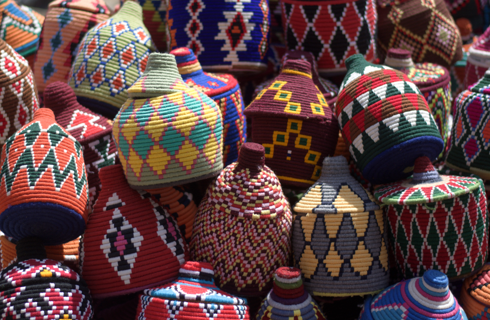 Colorful, intricately woven baskets are stacked together, displaying diverse geometric patterns and vibrant hues. The scene suggests a marketplace or craft fair showcasing traditional handmade artistry.