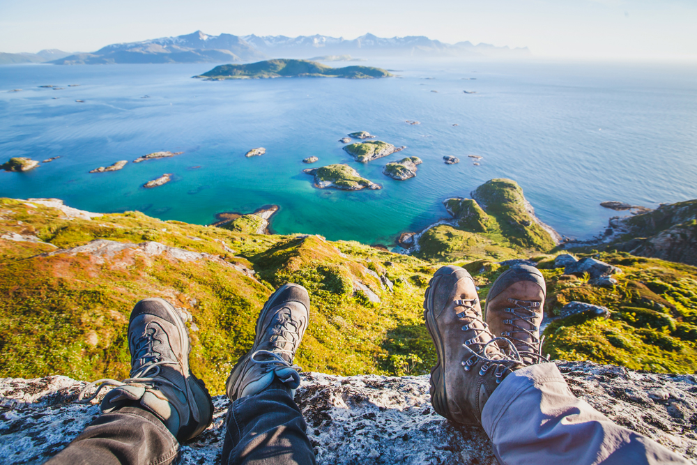 Two pairs of hiking boots rest on a rocky ledge overlooking a scenic coastal view with green islands, calm blue waters, and distant mountains under a clear sky.