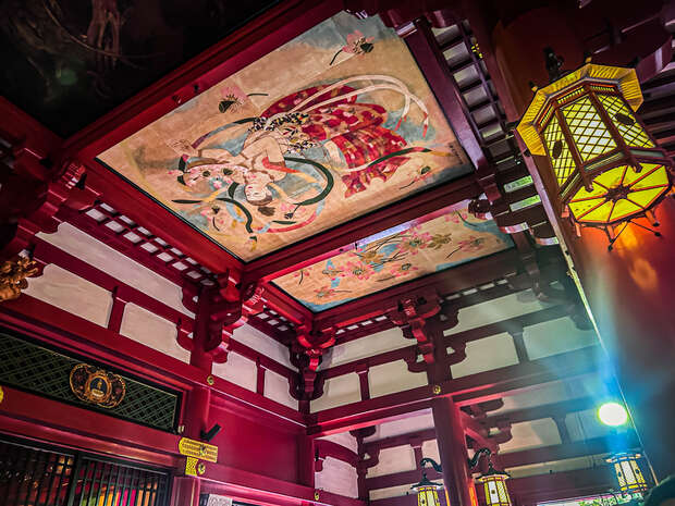 A vibrant ceiling painting depicts a mythological scene in a richly decorated temple with red beams. Surrounding lanterns add warmth, enhancing the traditional architectural features.
